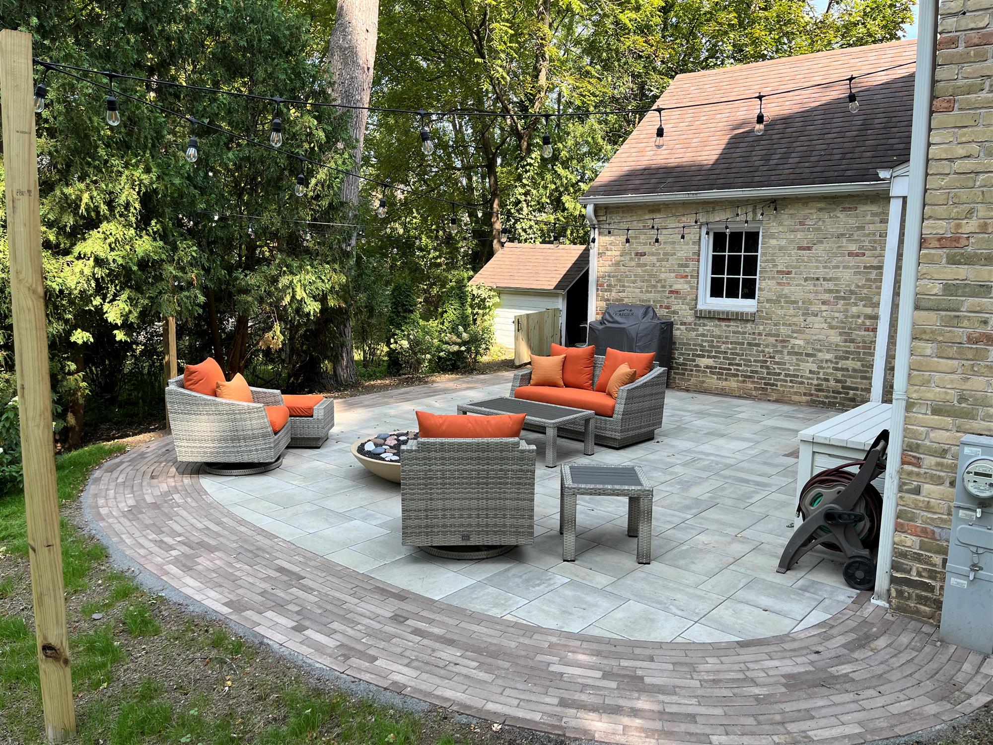 Brick patio connected to a house, surrounded by forest, with hanging lights, patio furniture, and a fire pit.