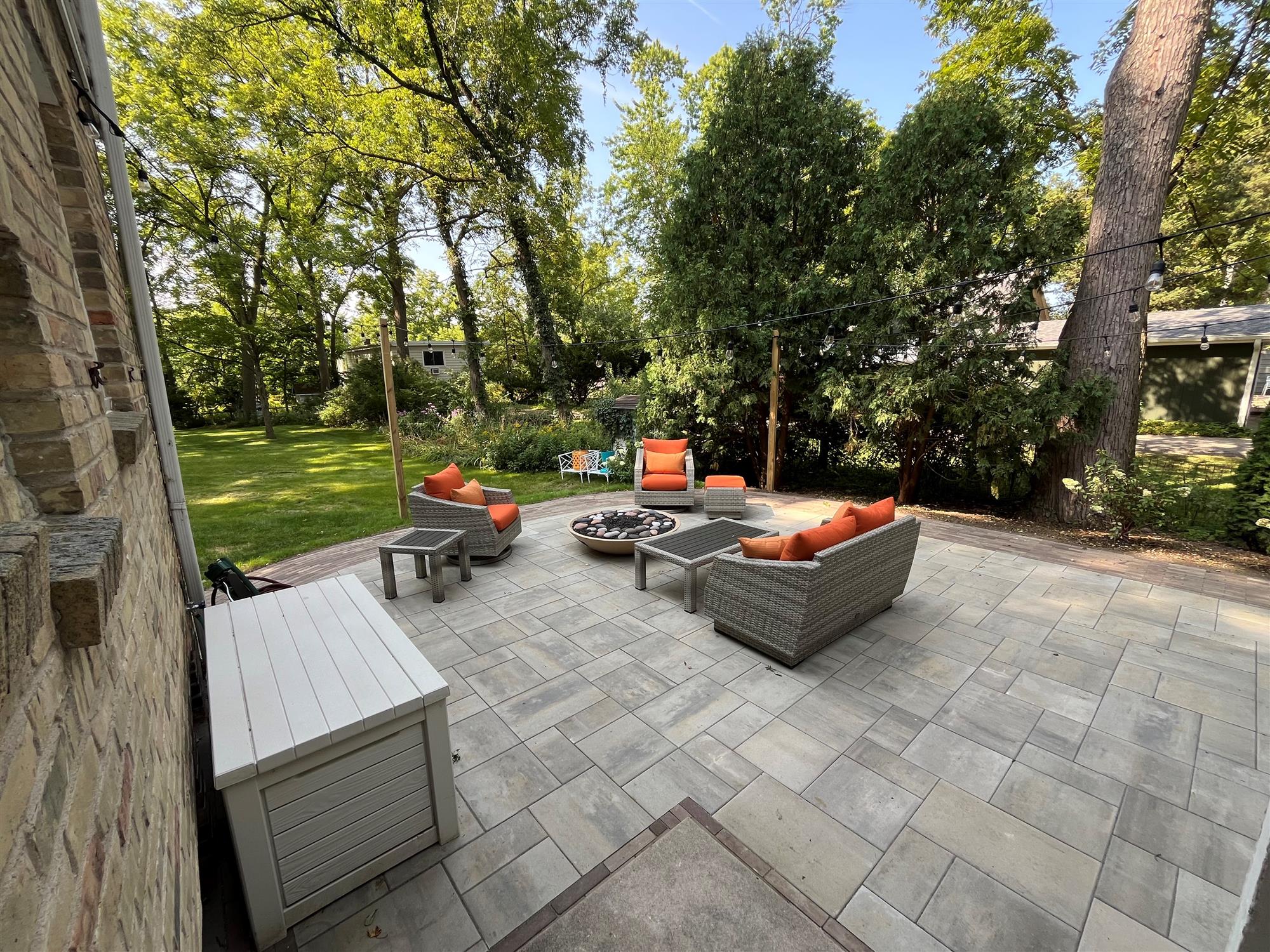 Patio with hanging lights, orange cushioned furniture, side tables, and a lush backyard surrounded by trees.