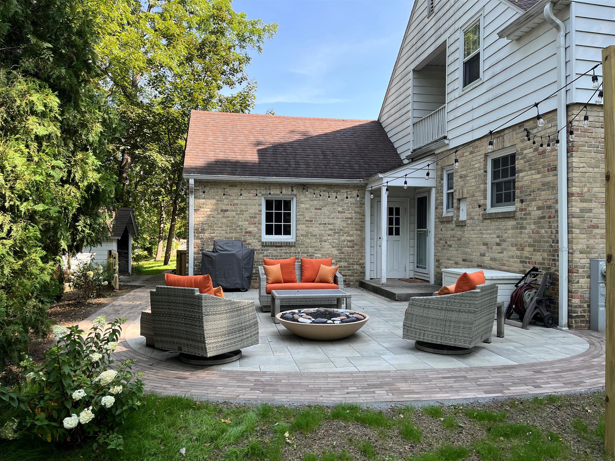 Secluded patio in the back corner of a house with orange cushions, small side tables, and a lush backyard surrounded by trees.